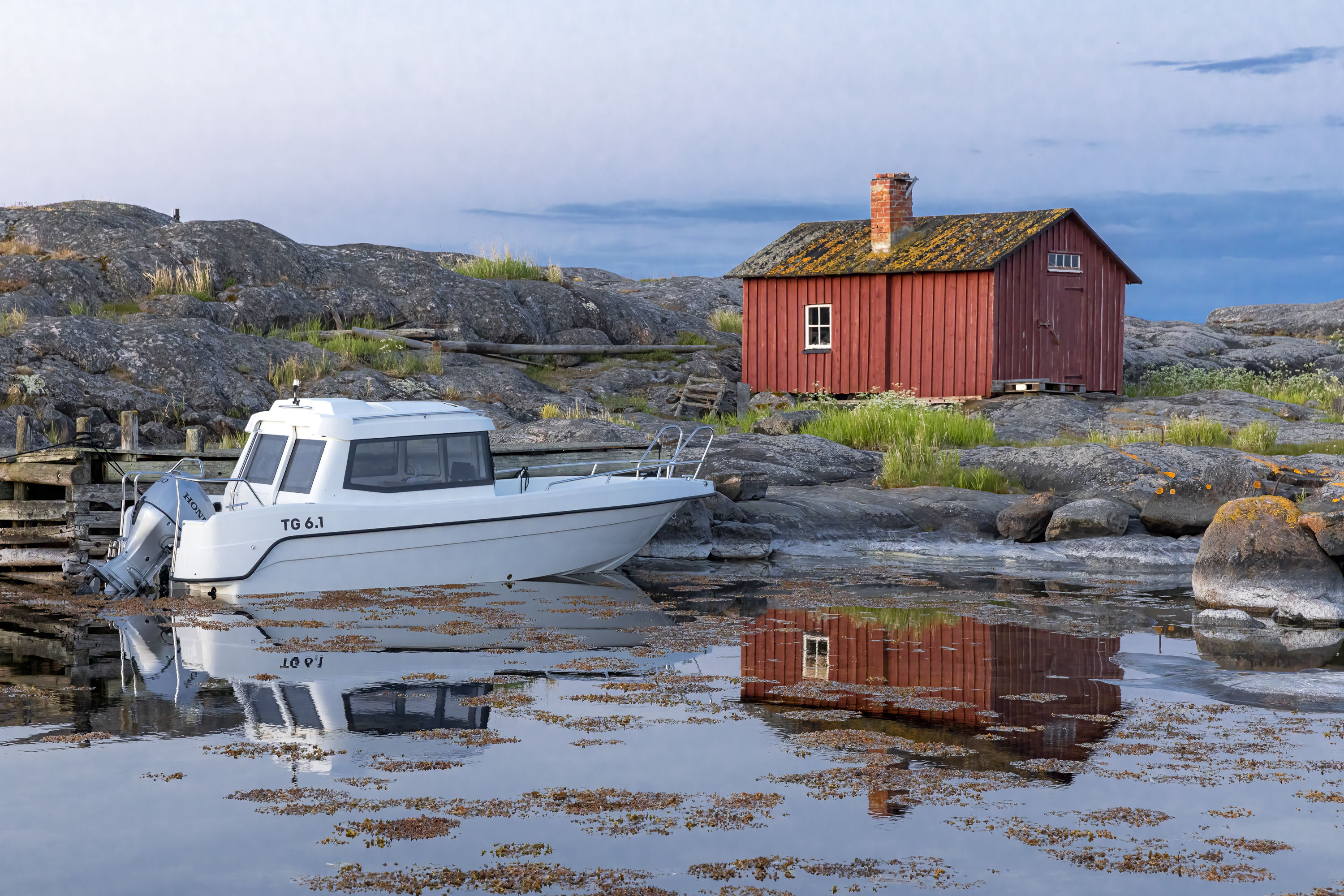 TG 6.1 kabinbåt står förtöjd vid gammal träbrygga i skärgården. Gammal röd fiskehytta i bakgrunden.