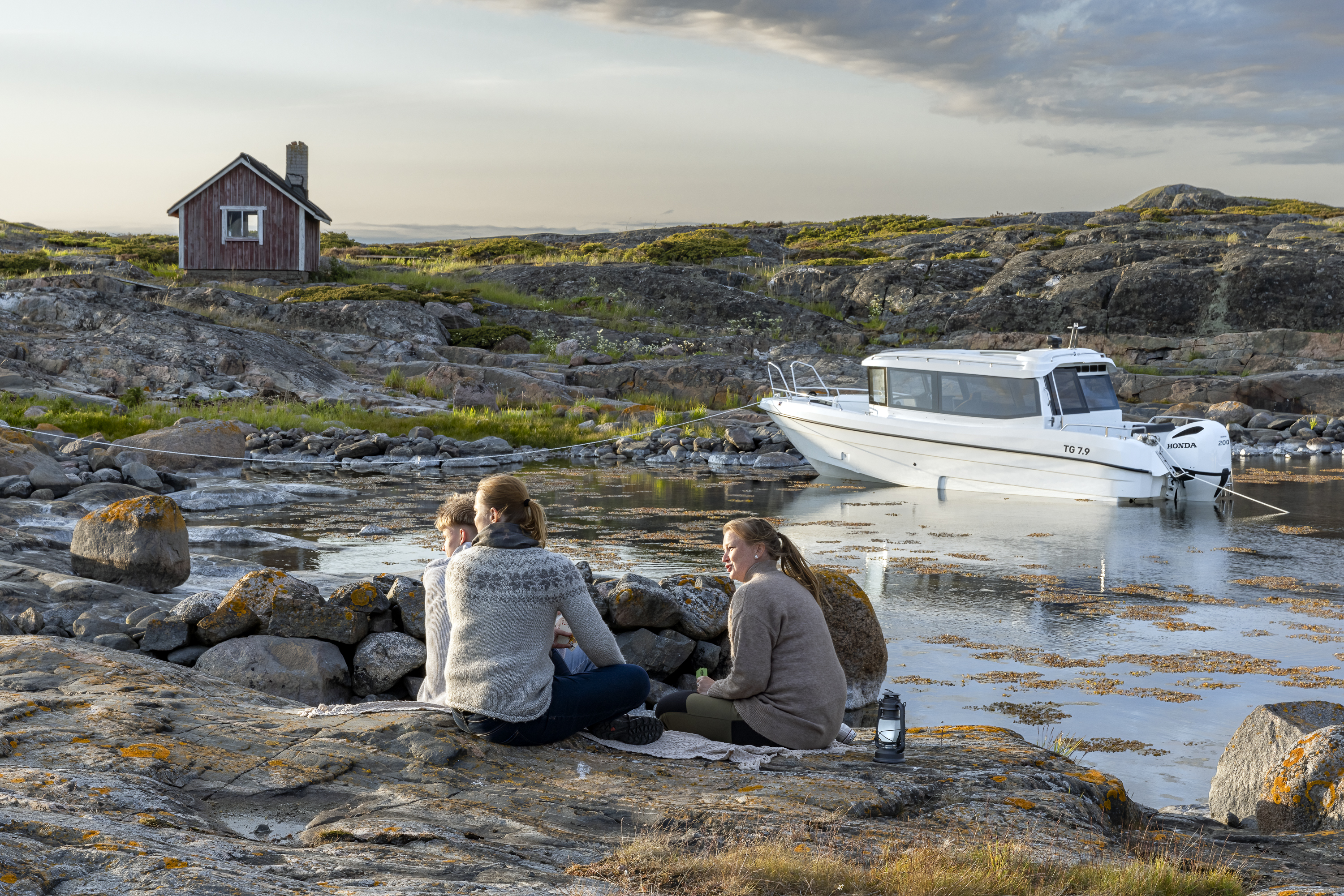 Kolme henkilöä viettää aikaa rantakalliolla ulkosaariston luonnonsatamassa, taustalla TG 7.9-hyttivene ja vanha kalamaja.